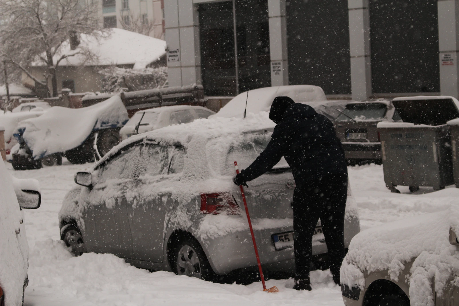 Person räumt bei Schneefall den Weg zwischen verschneiten Autos frei